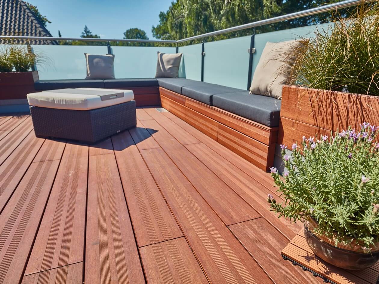 Vlondertips: onderhoud van een bamboe vlonder — Vlonderhoud Afgerond bamboe dakterras met warm roodbruine vlonderplanken, ingebouwde loungebank met kussens en plantenbakken achter een glazen balustrade — vlondertips van Vlonderhoud