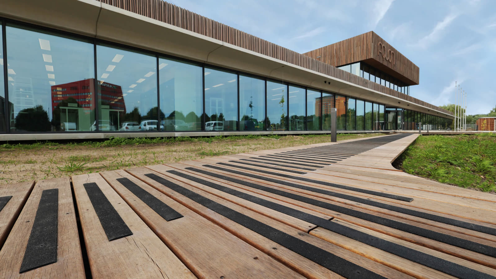 Antislip producten voor vlonders — Vlonderhoud Hardhouten vlonderpad met zwarte antislip-strips op elke plank langs een moderne kantoorgevel met glazen pui — antislip vlonder van Vlonderhoud